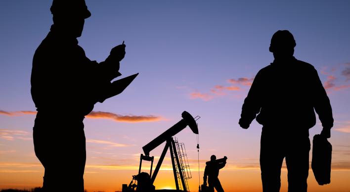 Silhouettes of 2 oil pump workers and an oil pump with sunset sky.